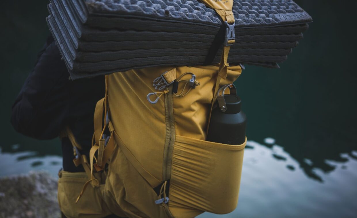 Yellow backpack with camping gear including a water bottle and folded mat near water.