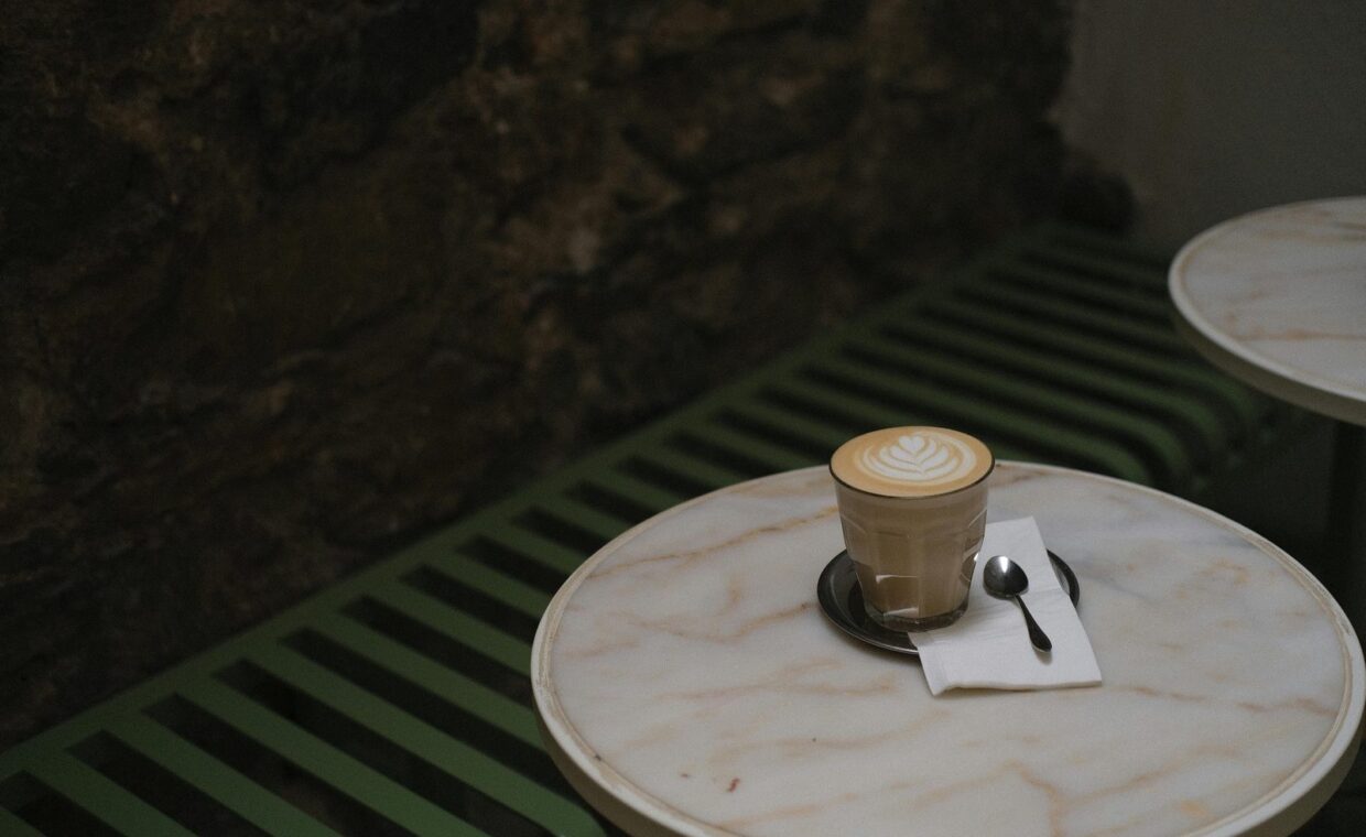 A beautifully crafted latte with delicate latte art sits on a marble table in a cozy café setting. The ambient lighting