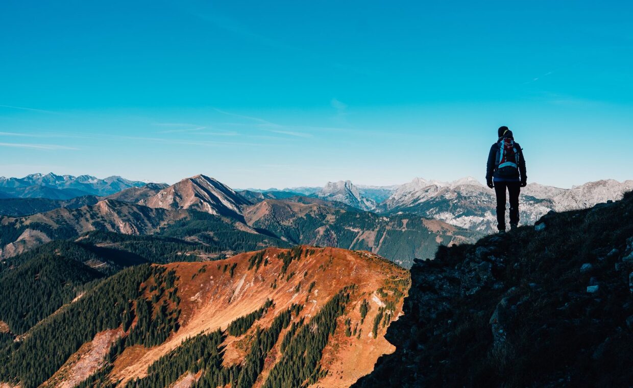 A lone hiker stands atop a mountain, taking in the breathtaking vista of mountain ranges under a clear blue sky.